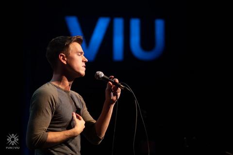 A man sings into a microphone with the letters VIU behind him